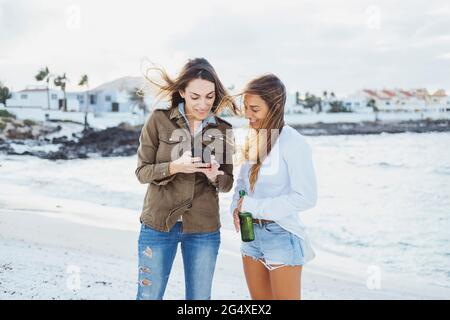 Donna mid adult che usa il telefono cellulare da un amico in piedi con una bottiglia di birra in spiaggia Foto Stock