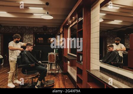 Giovane uomo che ottiene il taglio di capelli da barbiere maschio in salone durante la crisi COVID-19 Foto Stock