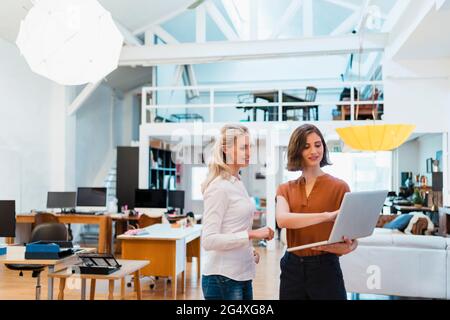 Donne professionisti che discutono sul laptop mentre si levano in piedi insieme in ufficio creativo Foto Stock