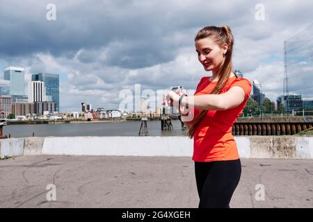 Donna sorridente in forma che controlla il tempo sul orologio da polso vicino al fiume in città Foto Stock