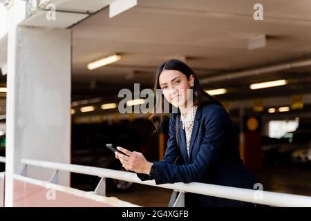 Donna d'affari sorridente con smartphone appoggiato sulla ringhiera nel parcheggio Foto Stock