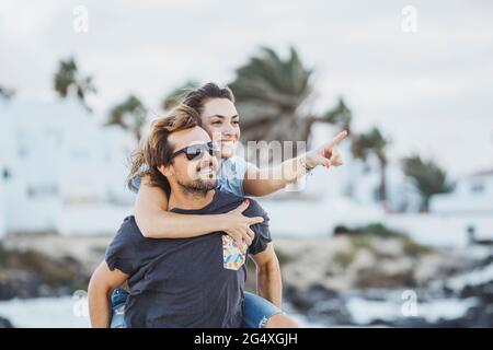 Donna sorridente che punta mentre piggybacking sull'uomo alla spiaggia Foto Stock