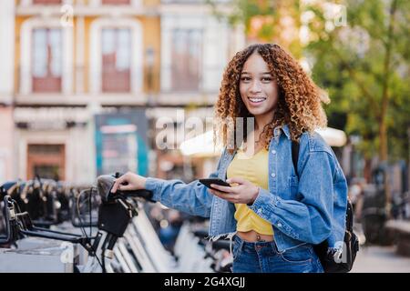 Donna sorridente con smartphone in piedi vicino alla stazione di parcheggio per biciclette Foto Stock