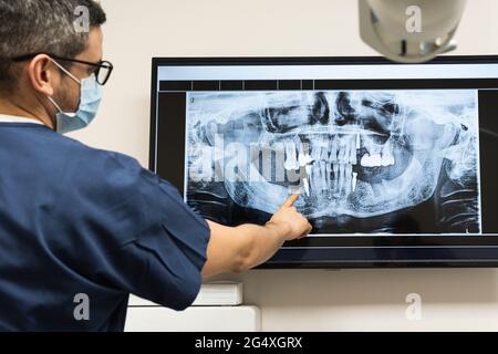 Dentista maschile indossando maschera protettiva per il viso esaminando radiografia dentale in clinica Foto Stock