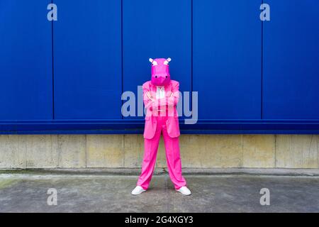 Donna che indossa un abito rosa vibrante e maschera ippopotamo in piedi davanti alla parete blu con le braccia incrociate Foto Stock