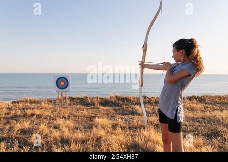 Archeress che mira a bersaglio mentre si trova in piedi su erba durante la giornata di sole Foto Stock