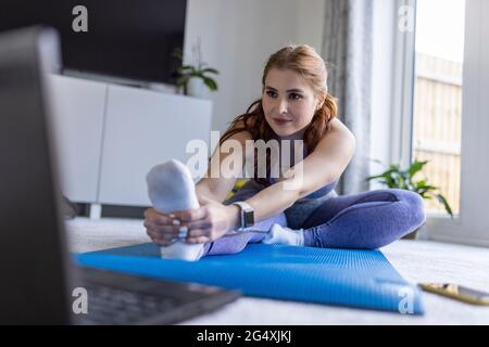 Donna redhead facendo esercizio stretching mentre imparando attraverso tutorial online a casa Foto Stock
