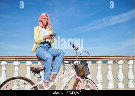 Giovane donna con bicicletta che usa lo smartphone mentre si siede sulla ringhiera a Promenade Foto Stock