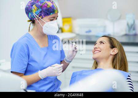 Dentista femminile che indossa una maschera protettiva che tiene in mano le attrezzature dentali mentre si parla con il paziente in clinica Foto Stock