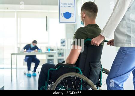 Operatore sanitario femminile che porta la paziente su sedia a rotelle al centro di vaccinazione COVID-19 Foto Stock