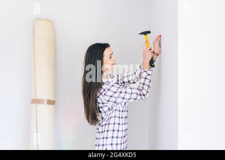 Donna sorridente che batte chiodo nel muro a casa Foto Stock