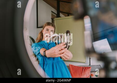 Ragazza adolescente che mostra diversi pennelli di make-up mentre vlogging a casa Foto Stock