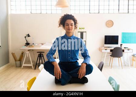 Donna d'affari che pratica yoga mentre si siede a gambe incrociate sulla scrivania in ufficio Foto Stock