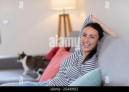 Giovane donna allegra con gatto tabby in background sul divano a casa Foto Stock