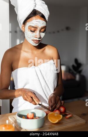 Donna con maschera facciale che indossa asciugamano mentre taglia la frutta in cucina a casa Foto Stock