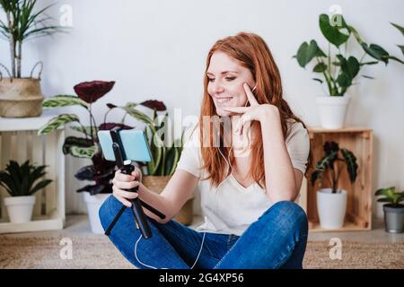 Bella donna sorridente durante la videochiamata con smartphone sul pavimento a casa Foto Stock