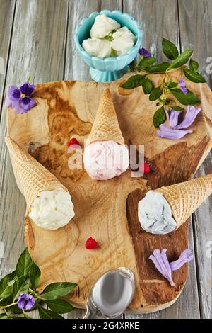 Gelato alla vaniglia, al mirtillo e al lampone in coni di waffle su tavola di legno con frutti di bosco e fiori Foto Stock
