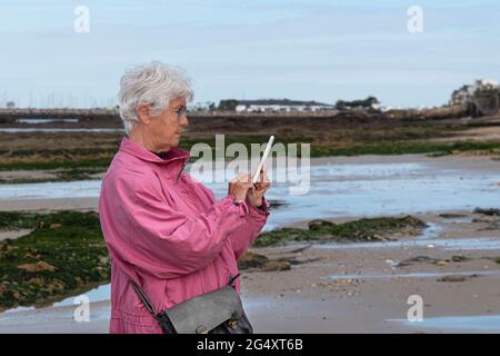 Una donna anziana scrive un messaggio di testo sul suo telefono sulla spiaggia in Bretagna Foto Stock