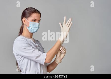 un medico in un cappotto bianco e una maschera medica mette guanti sterili in lattice bianco sulle sue mani per la procedura, spazio di copia Foto Stock