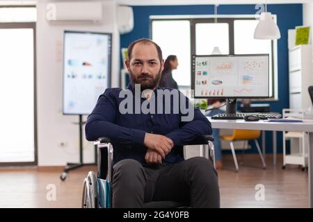 Triste uomo d'affari frustrato delle sue disabilità guardando la fotocamera seduto immobilizzato in sedia a rotelle in ufficio lavoro su pc con i dati, protesi informazioni finanziarie. Foto Stock