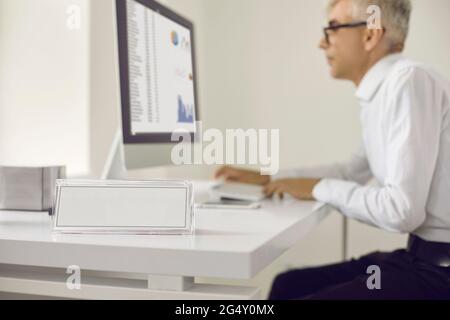 Primo piano della targhetta bianca vuota in ufficio sulla scrivania sullo sfondo di un uomo che lavora su un computer portatile. Foto Stock