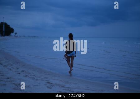 La giovane donna tailandese sta camminando attraverso la spiaggia della sera, a piedi nudi, con i suoi sandali in mano, un po 'di bassa marea Foto Stock