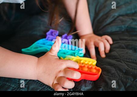 Bambina con giocattolo popit in colori arcobaleno. Giocattolo in silicone a forma di dino per ridurre lo stress. Bolle sensoriali fidget trendy. Bambino che gioca a casa Foto Stock