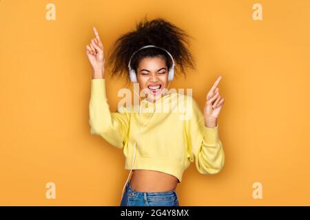 Gioiosa ragazza in cuffia, alza la mano in movimento danza e canta forte su sfondo giallo Foto Stock