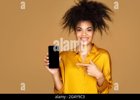 La ragazza molto contentata punta il dito sul telefono cellulare, consiglia qualcosa. Foto di ragazza afroamericana su sfondo beige Foto Stock