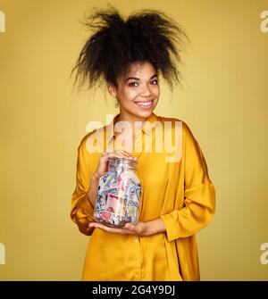 Sorridente ragazza mostra i suoi soldi di risparmio in vaso di vetro. Foto di ragazza afroamericana su sfondo giallo Foto Stock