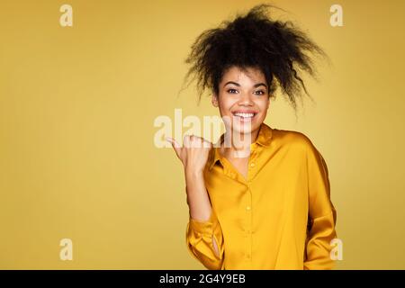 Sorridente ragazza punti da parte con il pollice, mostra sullo spazio di copia per il tuo testo. Foto di ragazza afroamericana su sfondo giallo Foto Stock