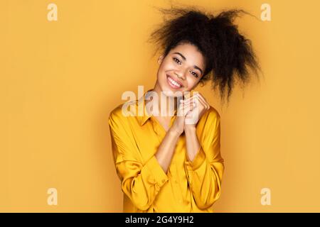 Bella ragazza sorridente tiene le mani pressate insieme sotto il mento, guarda qualcosa di carino. Foto di ragazza afroamericana su sfondo giallo Foto Stock