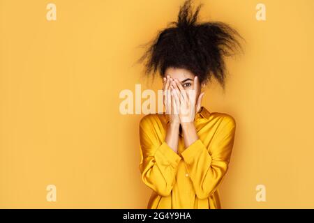 La ragazza spaventata sbirca attraverso le dita, copre faccia con entrambe le mani. Foto di ragazza afroamericana su sfondo giallo Foto Stock