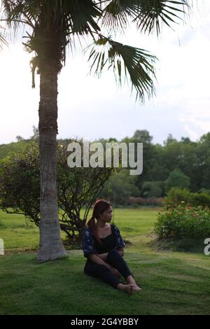 Una donna thailandese è seduta sotto la palma su un prato rifinito, che si affaccia su un parco durante l'ora d'oro Foto Stock