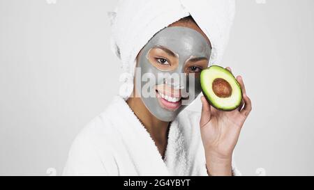 Donna afroamericana sorridente con maschera di argilla sul viso che tiene avocado isolato su grigio Foto Stock