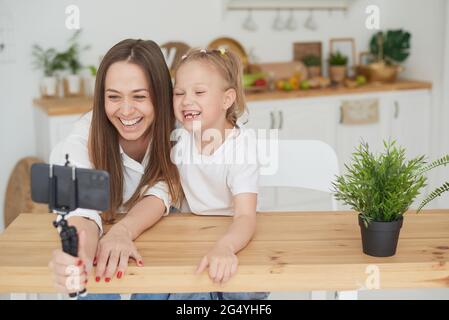 Una famiglia felice. Carina piccola figlia è seduta accanto a mamma in cucina, ridendo, divertendosi con uno smartphone, guardando un video divertente. Video com Foto Stock