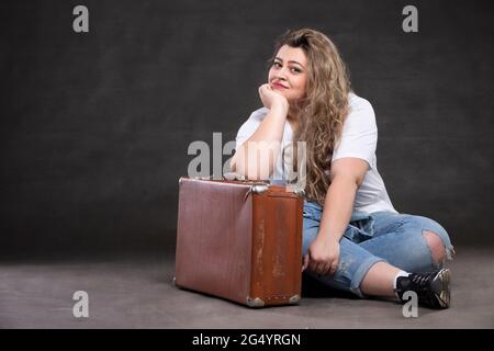 Bella donna grassa in denim vestiti con una vecchia valigia retrò su uno sfondo grigio. Modello ragazza più grande. Foto Stock