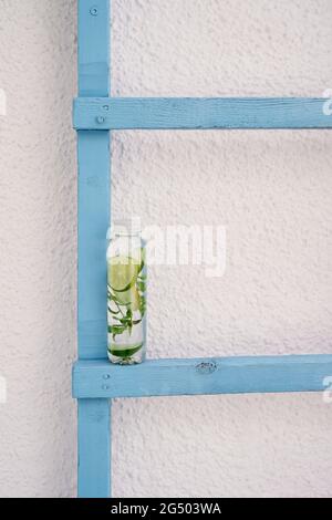 Una bottiglia d'acqua con fette di limone, lime e menta si trova su una scala in legno vicino al muro Foto Stock