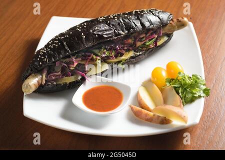Hot dog nero lungo con salsiccia di pollo fritta con insalata, su un piatto con salsa, pomodori e patate Foto Stock