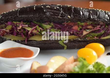 Hot dog nero lungo con salsiccia di pollo fritta con insalata, su un piatto con salsa, pomodori e patate, primo piano Foto Stock