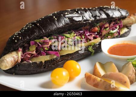 Hot dog nero lungo con salsiccia di pollo fritta con insalata, su un piatto con salsa, pomodori e patate, primo piano Foto Stock
