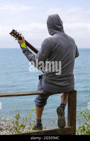 Giovane caucasico seduto sulla ringhiera sulla costa del mare e suonando la chitarra. Vista dal retro Foto Stock