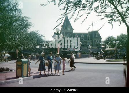 Disneyland, California, 1959. Vista sulla Piazza della Città e sulla stazione della strada principale. Un'auto trainata da cavalli Disneyland e il Disneyland Omnibus sono in attesa fuori dalla stazione. Quest'ultimo sta visualizzando il banner "Disneyland ‘59" che sta pubblicizzando le nuove aggiunte del residence per quell'anno. Un antico cannone è in mostra nella piazza, mentre i visitatori si rilassano sulle panchine e passano accanto. Un gruppo di tre Signore che accompagnano due ragazze giovani stanno per attraversare la strada mentre in conversazione. Foto Stock