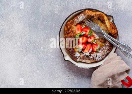 Frittella olandese per bambini con fragola fresca e cosparsa di zucchero a velo in polvere in padella rossa su fondo bianco della cucina. Vista dall'alto. Foto Stock