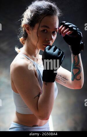Ritratto di una giovane donna che indossa guanti da boxe, guardando la fotocamera. Ha i suoi pugni in su davanti alla sua faccia ed è pronta a combattere. Foto Stock