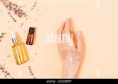 Mano femminile con campione applicato di maschera cosmetica in argilla accanto a bottiglie di olio essenziale di lavanda e olio nutriente su sfondo beige. Foto Stock