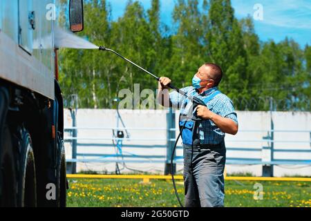 Autolavaggio per veicoli pesanti e cargo. Un uomo UN operaio del lavaggio dell'automobile in una maschera medica lava un camion con una pistola dell'acqua sulla strada in un giorno d'estate. Foto Stock
