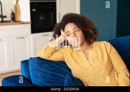 La giovane donna frustrata si siede sul divano a casa da sola, la donna africana sconvolsa si sente sola e misera, la donna disoccupata si preoccupa del futuro Foto Stock