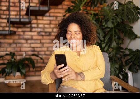 Gioiosa giovane afroamericana che usa uno smartphone seduto sulla poltrona di casa, sorridente donna nera ama chattare online, passare il tempo in social network Foto Stock