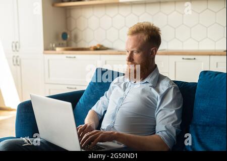 Uomo rosso focalizzato utilizzando il computer portatile per studiare o lavorare online, ragazzo freelance digitando seduto sul divano a casa, sviluppa nuovo progetto lavorando a distanza Foto Stock